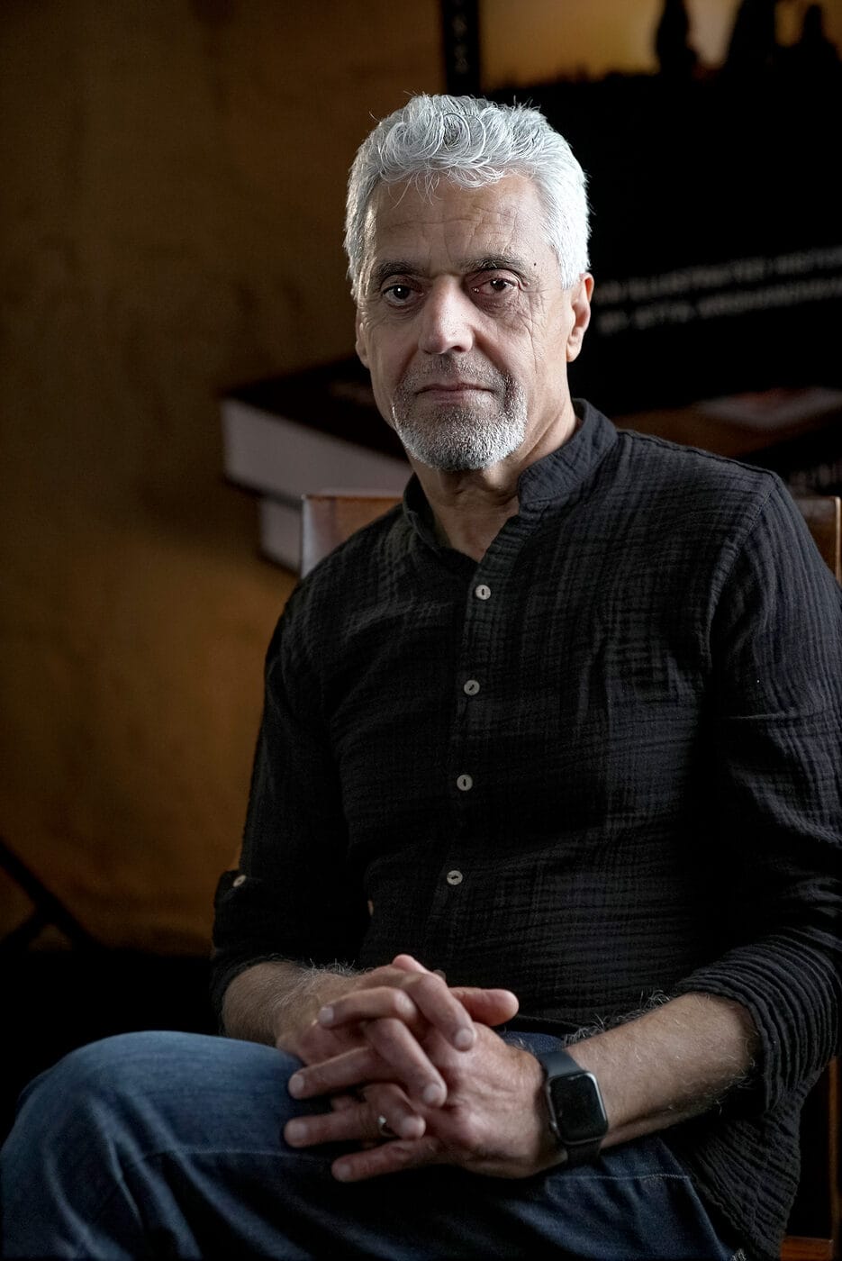 Atta Arghandiwal seated with his hands folded, in a warm, low-lit editorial portrait.